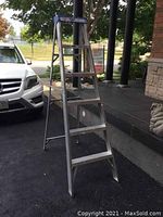 Photo of a silver aluminum folding step ladder approximately 6 feet tall standing on asphalt outdoors. It has a blue top platform and six wide steps visible. The ladder is set up in an A-frame position for standing use.