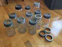 11 clear glass canning jars with blue-green glass sealers and metal screw-on rings, laid out on wooden floor. Four extra metal rings are separate in front.