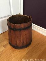 Photo of an antique wooden bucket with vertical slats and two black metal rings on hardwood flooring against white and dark walls.