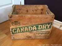 Photo of a rectangular wooden crate with natural wood color and aged condition. The front panel has a green painted Canada Dry logo and slogan.