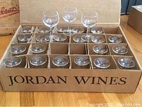 24 clear wine glasses in a cardboard box with dividers labeled Jordan Wines, arranged upright.