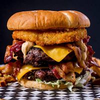 High-quality close-up photo of a double cheeseburger with bacon, cheddar cheese, lettuce, sauce, and a large onion ring on a bun, showcasing an example of World Burger's menu offerings.