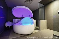Photo of the open sensory deprivation flotation therapy tank, white egg-shaped with lid elevated showing inside with blue light.