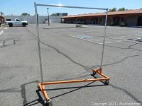 Full view photo of the heavy duty clothing rack with a chrome top bar and orange Z-style base with large caster wheels on asphalt in a parking lot.