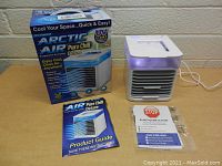 Arctic Air Pure Chill Deluxe personal fan cooler plugged in and lit with purple light, placed next to original box and product guide on a brick background.