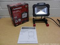 Photo showing Husky 2000 Lumen LED Work Light alongside its original box and instruction manual on a beige surface.