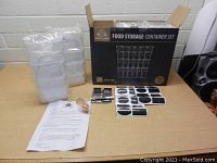Photo showing the box labeled 'Food Storage Container Set' with plastic containers stacked beside it, chalkboard labels, marker, spoon set, and instruction sheet on the table.
