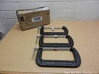 Three large Husky heavy duty 8 inch C clamps displayed on a table, with the original packaging box behind them.