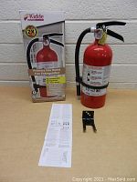 Full view of Kidde fire extinguisher next to original box, mounting bracket, and instruction manual laid flat on a table.