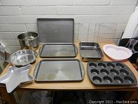 Photo showing all baking pans, sheets and racks on a table including star-shaped pan, heart pan, muffin tin, cookie sheets, square pan, cooling racks, metal bowl and colander.