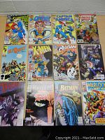 Top-down photo showing 12 assorted comic books neatly arranged in three rows on a wooden table against a grey wall. Comics include Superman, Quasar, Captain America, Justice League, ElfQuest, X-Men, Robin, Ultimate Captain America, Thor, Wolverine, Batman, and Warlock.