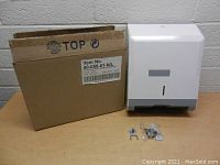 White wall mount paper towel dispenser next to its original cardboard box. Small plastic bag with mounting screws and hardware is in front of the dispenser.