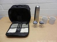 Front view of travel coffee set components displayed against a white brick wall background. Shows the carry case open with utensils and napkins, thermos, mugs, and sealed containers.