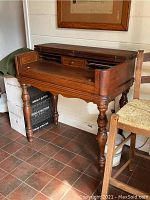 Angle view of antique wooden desk showing spindle legs, hinged top closed, under a framed picture.