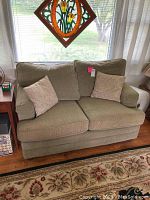 Front view of the Bassett two-seat green sofa with two beige decorative pillows on a floral rug near a window.