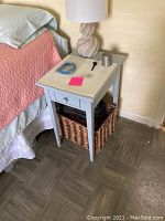 Bedside table next to bed with lamp, small items on top, drawer and basket below