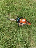 Stihl 021 orange chainsaw lying on grass, showing left side view with chain bar and handle