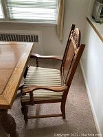 Captain chair next to dining table showing carved wooden back, striped upholstered seat, and wooden armrests.
