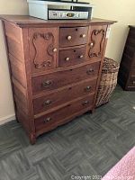 Side angled view of vintage wooden dresser showing detailed carved cabinet panels, different drawer sizes, and metal and wooden knobs/handles. Shows general condition and wood grain.