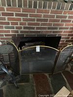 Front view of a three-panel metal fireplace screen with brass finish standing in front of a brick fireplace.