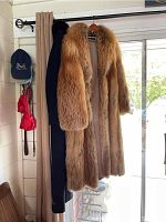 Full view of two full-length coats hanging on curtain rod by window: a reddish-brown fox fur coat and a black wool coat with fur collar.