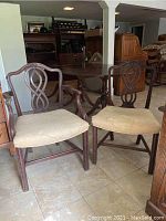 Photo showing a pair of mahogany dining chairs: one captain's chair with armrests and one standard side chair with more ornate backrest. Upholstery is visibly stained and worn.