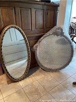 Both vintage mirrors shown leaning against wood furniture on tiled floor. Oval mirror is uncovered; round mirror is wrapped in bubble wrap.