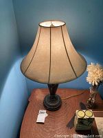 Full view of lamp showing beige bell-shaped fabric shade, lamp height, and its placement on table near wall.
