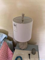 Table lamp with beige swirl-style base and white fabric drum shade, viewed from front on bedside table with some small items around.