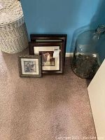 Three framed art pieces leaning against a blue wall, two larger floral designs in wood-like frames and a smaller framed piece with a detail-rich design