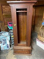 Front view of tall wooden weapon cabinet with missing glass door panel showing the single drawer at the bottom.