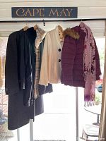Four outerwear pieces hanging on rack, including men's gray cashmere coat, Burberry plaid raincoat, cream jacket, maroon parka with purple scarf.