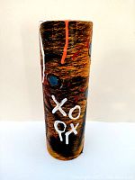 Front view of the spray can shaped sculpture showing black scribbled patterns, blue circles, white XO markings and orange paint drips on a bronze textured background.