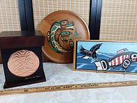 Photo showing three Indigenous art pieces: wooden box with carved front, wooden tray with painted design, and flat wood artwork with birds.
