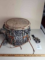 Front view of the African drum and the accompanying music shaker on a table. The drum features black and white spotted fabric on its sides and leather ties. The shaker has several metal jingles on a strap.