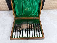 Photo showing the cutlery set arranged inside the wooden box with green velvet base and mother of pearl handles visible.