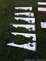 Six white-painted antique wooden porch brackets laid on grass showing decorative carvings and aged paint, some with chip damage exposing wood.