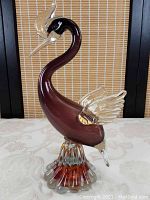 Full view of art glass bird sculpture with clear wings and gold flecks, burgundy body, and clear base.