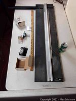 Top-down photo showing full large mat cutter on black metal base, aluminum cutting bar, two boxes with cutter heads, wooden ruler, and soda can for scale.