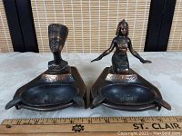 Pair of Egyptian vintage copper ashtrays featuring detailed figure sculptures on triangular bases with oval ashtray bowls, placed next to a ruler showing size.