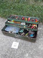 Full view of open green metal fishing tackle box showing two trays filled with various fishing lures and reels in bottom compartment.