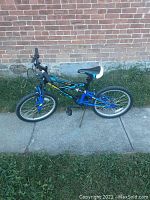 Full side view of the Momentum Supercycle kids bike leaning against a brick wall. Shows overall design, frame color, tires, and seat.