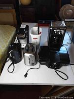 Full lot overview showing Keurig coffee maker, Bella coffee brewer, Hamilton Beach grill, toaster, kettle, and miscellaneous items on a white table.