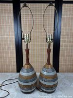 Pair of mid-century lamps showing ceramic bases with horizontal decorative bands and polished wooden necks, without shades, cords visible.