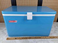 Front view of the blue Coleman cooler showing latch and brand label with ruler beneath showing size.