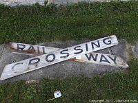 Two aged white wooden railway crossing sign boards with black lettering placed on grass and concrete.