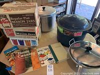 Three stacked boxes containing Mason jars: Kerr Regular Pint Mason Jars, Ball Regular Pint Mason Jars, and Ball Jelly Jars. Two metal stock pots with racks inside and a jar grabber tool on table. Five assorted canning cookbooks laid out.