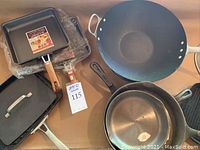 Top view of various skillets including cast iron and nonstick, some still wrapped in plastic, arranged on a table.