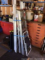 Photo showing two step ladders and a folding step stool bundled together under garage workshop shelves with tools and paint cans in background.