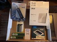 Overview photo showing all items in the lot: ASUS ZenDrive external DVD writer, 4 port USB 3.0 hub, two sticks of RAM, two electronic lighters, Crucial MX500 SSD, and computer hardware parts.